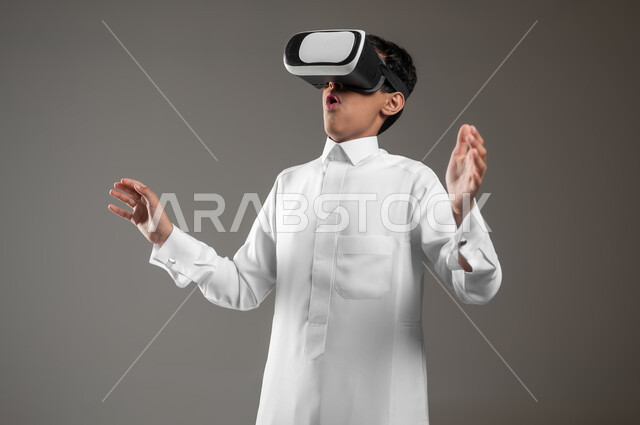 محاكاة العالم الخيالي والتفاعل مع الفيلم، تعابير وايماءات تدل على الاندماج والدهشة, بورتريه مقرب لرفف لولد عربي خليجي سعودي متفاجئ يرتدي الثوب التقليدي يقوم بمشاهدة أفلام عبر نظارات الواقع الافتراضي ثلاثية الأبعاد, خلفيه رمادية، محتوى حساس، استخدام حساس