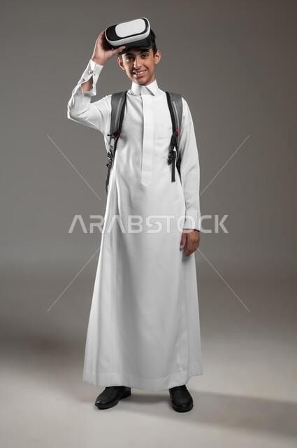 النظر إلى الكاميرا بتعابير السرور، بورتريه مقرب لطالب عربي خليجي سعودي مبتسم يرتدي الثوب التقليدي وحقيبة الظهر المدرسية يضع نظارة الفي آر، نشاط دراسي باستعمال الواقع الافتراضي، صورة الجسم بالكامل، خلفية رمادية، محتوى حساس، استخدام حساس