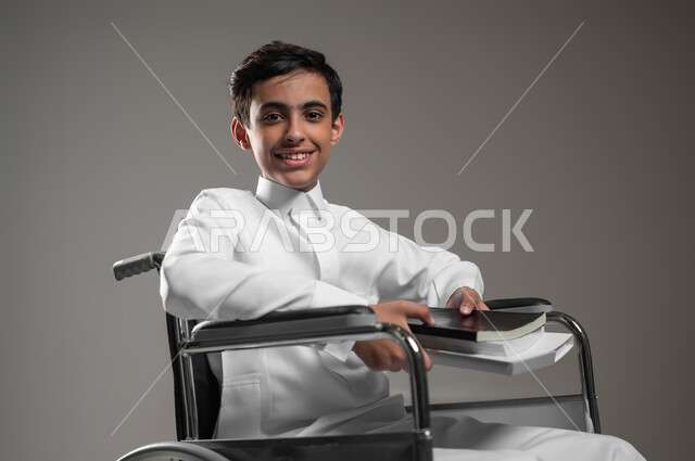 الاهتمام بتعليم ذوي الاحتياجات الخاصة, دمج المعاقين في المدارس السعودية, تنمية القدرات العقلية والفكرية لذوي الهمم, بورتريه مقرب لولد عربي خليجي سعودي مبتسم يرتدي الثوب التقليدي يجلس على الكرسي المتحرك يحمل الكتب الدراسية, خلفية رمادية، محتوى حساس، استخدام حساس