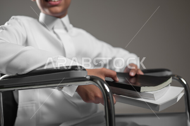 دمج المعاقين في المدارس السعودية, الاهتمام بتعليم ذوي الاحتياجات الخاصة, تنمية القدرات العقلية والفكرية لذوي الهمم, بورتريه مقرب لولد عربي خليجي سعودي مبتسم يرتدي الثوب التقليدي يجلس على الكرسي المتحرك يحمل الكتب الدراسية, خلفية رمادية، محتوى حساس، استخدام حساس