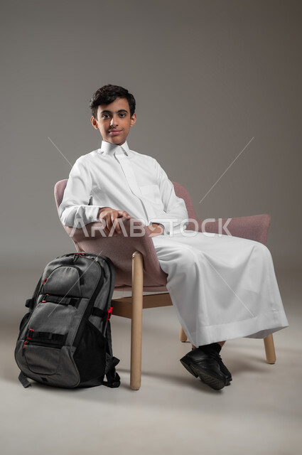 النظر الى الكاميرا بتعابير السرور، التعليم في المملكة العربية السعودية، بورتريه لطالب عربي خليجي سعودي يرتدي الثوب التقليدي يجلس على كرسي مريح بجانب حقيبة الظهر المدرسية، بداية العام الدراسي الجديد, مفهوم الجلوس والراحة، خلفية رمادية، محتوى حساس، استخدام حساس