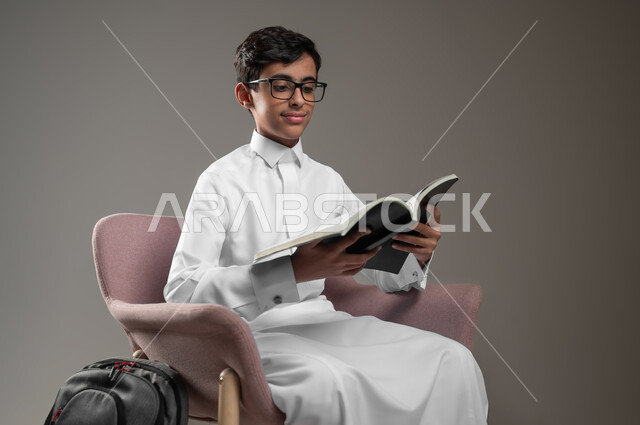 الاجتهاد وتحضير الدروس, متابعة وحل الواجبات اليومية، تطوير المناهج التعليمية في السعودية, بورتريه لولد عربي سعودي خليجي مبتسم يرتدي الثوب التقليدي يجلس على الكرسي يقوم بقراءة الكتب, بداية العام الدراسي الجديد, خلفية رمادية، محتوى حساس، استخدام حساس