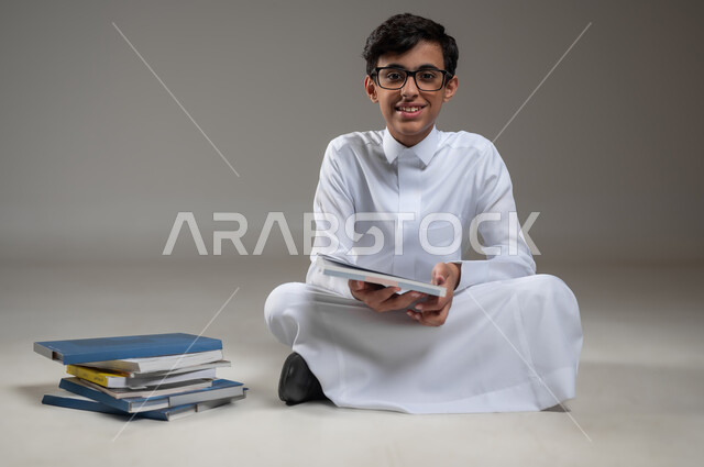 متابعة الدروس والواجبات اليومية، تطوير المناهج التعليمية في السعودية, الاجتهاد وتحضير الدروس, بورتريه مقرب لولد عربي سعودي خليجي مبتسم يرتدي الثوب التقليدي يجلس على الأرض يقوم بقراءة الكتب المدرسية, بداية العام الدراسي الجديد, خلفية رمادية، محتوى حساس، استخدام حساس