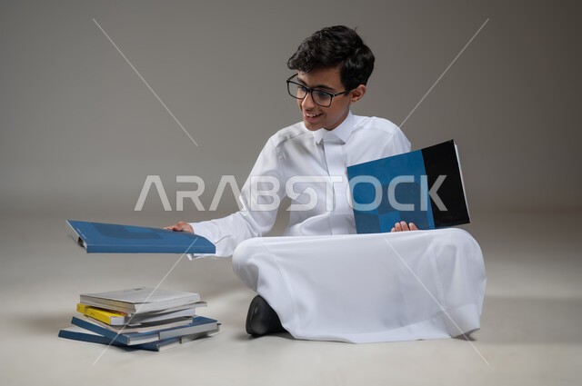 متابعة الدروس والواجبات اليومية، تطوير المناهج التعليمية في السعودية, الاجتهاد وتحضير الدروس, بورتريه مقرب لولد عربي سعودي خليجي مبتسم يرتدي الثوب التقليدي يجلس على الأرض يقوم بقراءة الكتب المدرسية, بداية العام الدراسي الجديد, خلفية رمادية، محتوى حساس،استخدام حساس
