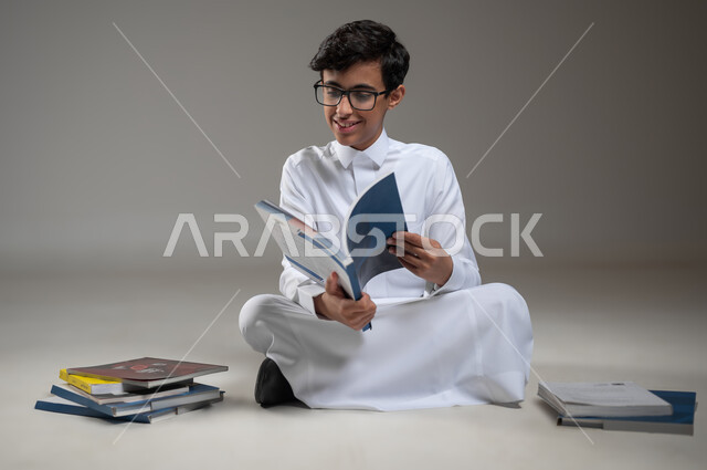 بداية العام الدراسي الجديد, متابعة الدروس والواجبات اليومية، تطوير المناهج التعليمية في السعودية, الاجتهاد وتحضير الدروس, بورتريه مقرب لولد عربي سعودي خليجي مبتسم يرتدي الثوب التقليدي يجلس على الأرض يقوم بقراءة الكتب المدرسية, خلفية رمادية، محتوى حساس، استخدام حساس