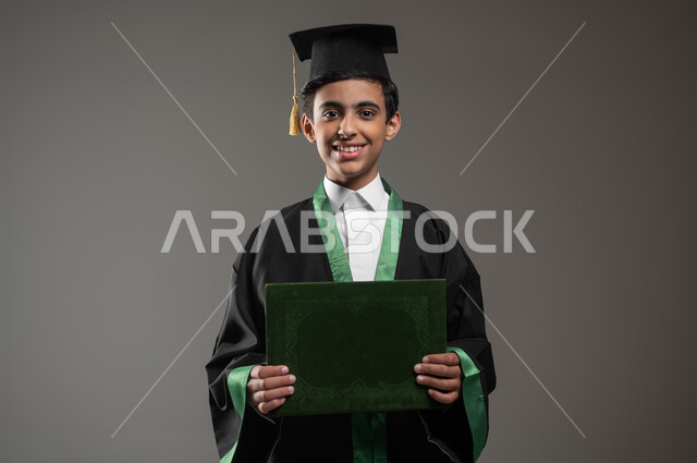 الاحتفال بالنجاح وانتهاء العام الدراسي، مفهوم التفوق والتخرج من المدرسة, النظر الى الكاميرا بتعابير وايماءات الفخر والسعاده, التعليم الأكاديمي في السعوديه, بورتريه مقرب لطالب عربي سعودي خليجي مبتسم يرتدي عباءه وقبعة التخرج يمسك بيده الشهادة، خلفية رمادية، محتوى حساس، استخدام حساس