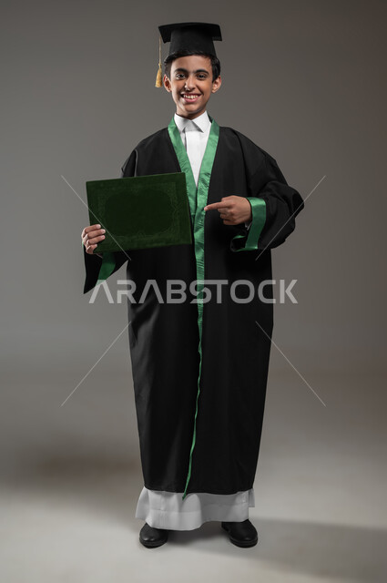 النظر الى الكاميرا بتعابير الفخر والسعاده, الاحتفال بالنجاح وانتهاء العام الدراسي، التعليم الأكاديمي في السعوديه, بورتريه لطالب عربي سعودي خليجي مبتسم يرتدي عباءه وقبعة التخرج يشير الى الشهادة، مفهوم التفوق والتخرج من المدرسة, صورة الجسم بالكامل، خلفية رمادية، محتوى حساس، استخدام حساس