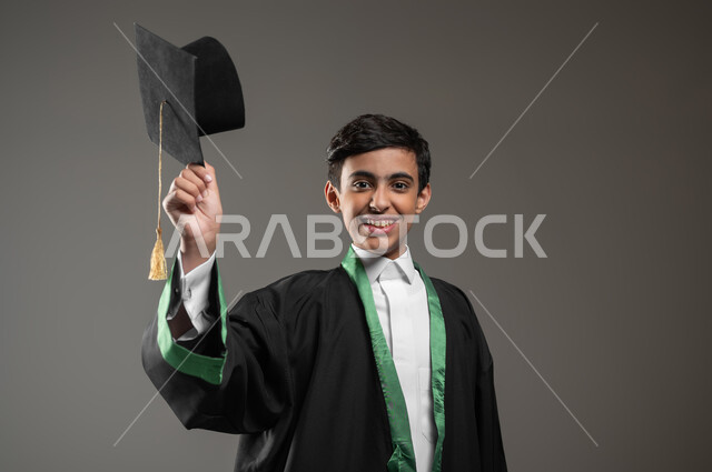 رفع القبعة عاليا بتعابير وايماءات الفخر والسعاده, التعليم الأكاديمي في السعوديه, بورتريه مقرب لطالب عربي سعودي خليجي مبتسم يرتدي زي التخرج يحتفل بالنجاح وانتهاء العام الدراسي، مفهوم التفوق والتخرج من المدرسة، خلفية رمادية،محتوى حساس، استخدام حساس
