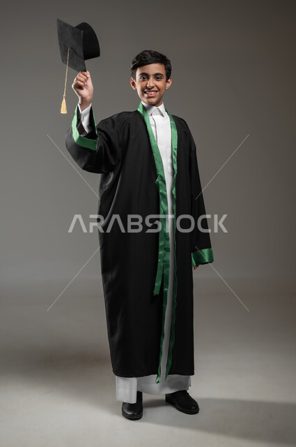 رفع القبعة عاليا بتعابير وايماءات الفخر والسعاده, التعليم الأكاديمي في السعوديه, بورتريه لطالب عربي سعودي خليجي مبتسم يرتدي زي التخرج يحتفل بالنجاح وانتهاء العام الدراسي، مفهوم التفوق والتخرج من المدرسة، صورة الجسم بالطول الكامل، خلفية رمادية، محتوى حساس، استخدام حساس