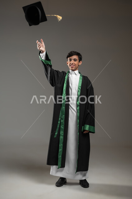 رمي القبعة عاليا بتعابير وايماءات الفخر والسعاده, التعليم الأكاديمي في السعوديه, بورتريه لطالب عربي سعودي خليجي مبتسم يرتدي زي التخرج يحتفل بالنجاح وانتهاء العام الدراسي، مفهوم التفوق والتخرج من المدرسة، صورة الجسم بالطول الكامل، خلفية رمادية، محتوى حساس، استخدام حساس
