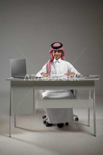 ادخال التقنية والتكنولوجيا بالتعليم، متابعة منصات التعليم الالكتروني المتاحة عبر الانترنت بإستخدام جهاز الحاسب المحمول، بورتريه لولد عربي خليجي سعودي يرتدي الشماغ والثوب التقليدي يقوم بحل الواجبات المنزليه عبر اللابتوب، خلفية رمادية، محتوى حساس، استخدام حساس