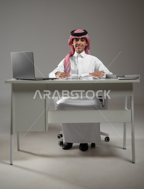 متابعة منصات التعليم الالكتروني المتاحة عبر الانترنت بإستخدام جهاز الحاسب المحمول، ادخال التقنية والتكنولوجيا بالتعليم، بورتريه لولد عربي خليجي سعودي يرتدي الشماغ والثوب التقليدي يقوم بحل الواجبات المنزليه عبر اللابتوب، خلفية رمادية، محتوى حساس، استخدام حساس