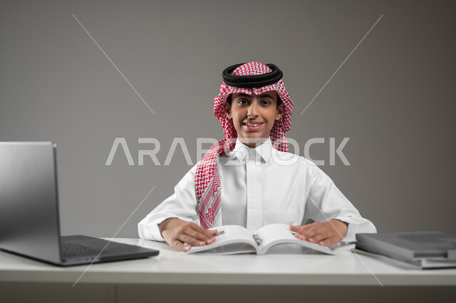 حل الواجبات المنزليه عبر اللابتوب، ادخال التقنية والتكنولوجيا بالتعليم، بورتريه مقرب لولد عربي خليجي سعودي يرتدي الشماغ والثوب التقليدي يقوم بمتابعة منصات التعليم الالكتروني المتاحة عبر الانترنت بإستخدام جهاز الحاسب المحمول، خلفية رمادية،محتوى حساس، استخدام حساس