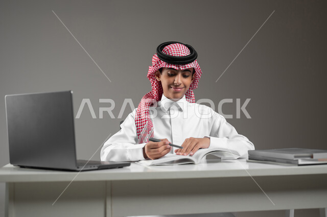 حل الواجبات المنزليه عبر اللابتوب، ادخال التقنية والتكنولوجيا بالتعليم، بورتريه مقرب لولد عربي خليجي سعودي يرتدي الشماغ والثوب التقليدي يقوم بمتابعة منصات التعليم الالكتروني المتاحة عبر الانترنت بإستخدام جهاز الحاسب المحمول، خلفية رمادية، محتوى حساس، استخدام حساس