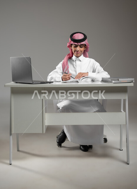 ادخال التقنية والتكنولوجيا بالتعليم، متابعة منصات التعليم الالكتروني المتاحة عبر الانترنت بإستخدام جهاز الحاسب المحمول، بورتريه لولد عربي خليجي سعودي يرتدي الشماغ والثوب التقليدي يقوم بحل الواجبات المنزليه عبر اللابتوب، خلفية رمادية،محتوى حساس، استخدام حساس