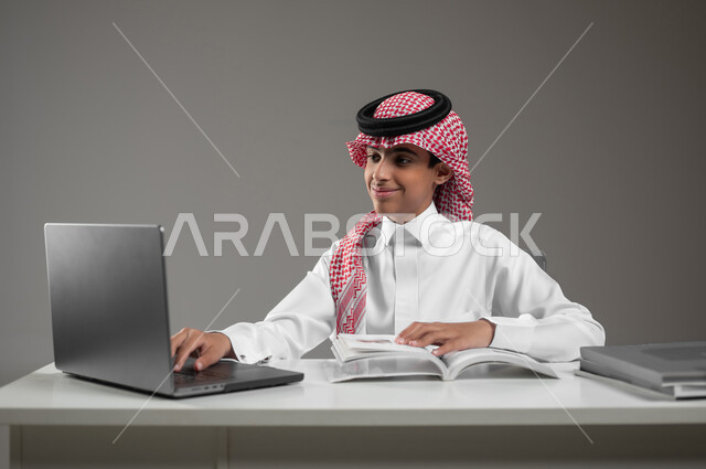 حل الواجبات المنزليه عبر اللابتوب، ادخال التقنية والتكنولوجيا بالتعليم، بورتريه مقرب لولد عربي خليجي سعودي يرتدي الشماغ والثوب التقليدي يقوم بمتابعة منصات التعليم الالكتروني المتاحة عبر الانترنت بإستخدام جهاز الحاسب المحمول، خلفية رمادية، محتوى حساس، استخدام حساس