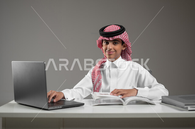 حل الواجبات المنزليه عبر اللابتوب، ادخال التقنية والتكنولوجيا بالتعليم، بورتريه مقرب لولد عربي خليجي سعودي يرتدي الشماغ والثوب التقليدي يقوم بمتابعة منصات التعليم الالكتروني المتاحة عبر الانترنت بإستخدام جهاز الحاسب المحمول، خلفية رمادية، محتوى حساس، استخدام حساس