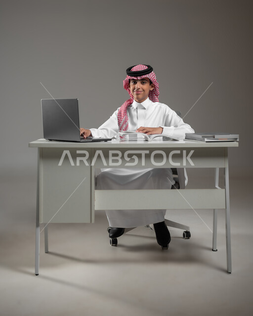 Following e-learning platforms available online using a laptop, introducing technology and technology into education, portrait of a Saudi Gulf Arab boy wearing a shemagh and traditional thobe, doing homework via the laptop, gray background, sensitive content, sensitive use