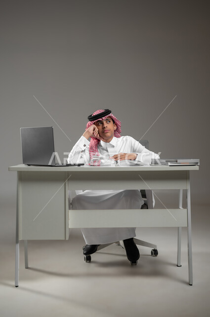 إيماءات التفكير بالمستقبل وبناءه, متابعة الدروس والدورات التعليمية عن بعد باستخدام اللابتوب, بورتريه لولد عربي خليجي سعودي يرتدي الشماغ والثوب التقليدي يجلس امام الكتب والحاسب المحمول, استعمال الأجهزة التقنية في التعلم, الشعور بالملل والضجر، خلفية رمادية،محتوى حساس، استخدام حساس