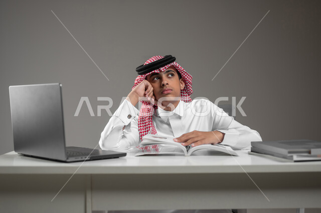 الشعور بالملل والضجر، إيماءات التفكير بالمستقبل وبناءه, متابعة الدروس والدورات التعليمية عن بعد باستخدام اللابتوب, بورتريه مقرب لولد عربي خليجي سعودي يرتدي الشماغ والثوب التقليدي يجلس امام الكتب والحاسب المحمول, استعمال الأجهزة التقنية في التعلم، خلفية رمادية، محتوى حساس، استخدام حساس