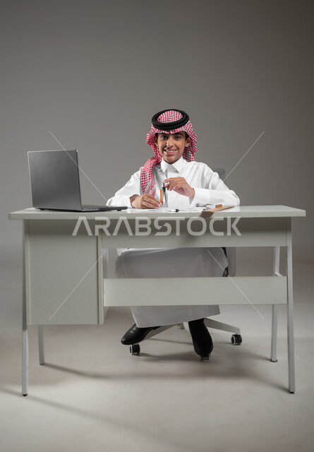 رسم مخطط تصميم هندسي، اكتساب المعرفة والخبرة بإيماءات الاستمتاع والسرور، إستخدام الأجهزة التقنية الحديثة في المجالات العلمية، بورتريه لولد عربي خليجي سعودي يرتدي الشماغ والثوب التقليدي يجلس امام حاسوبه يقوم باستعمال الأدوات والمعدات الهندسية، خلفية رمادية، محتوى حساس، استخدام حساس