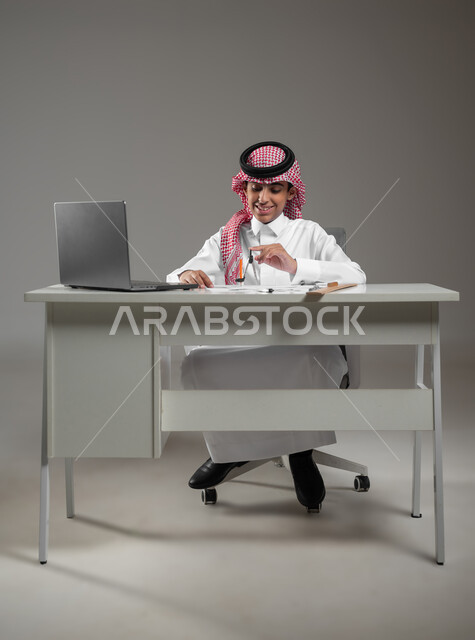 Gaining knowledge and experience with gestures of precision and focus, drawing an engineering design diagram, using modern technical devices in scientific fields, a portrait of a Saudi Arabian Gulf boy wearing a shemagh and traditional dress sitting in front of his computer using engineering tools and equipment, gray background, sensitive content, sensitive use