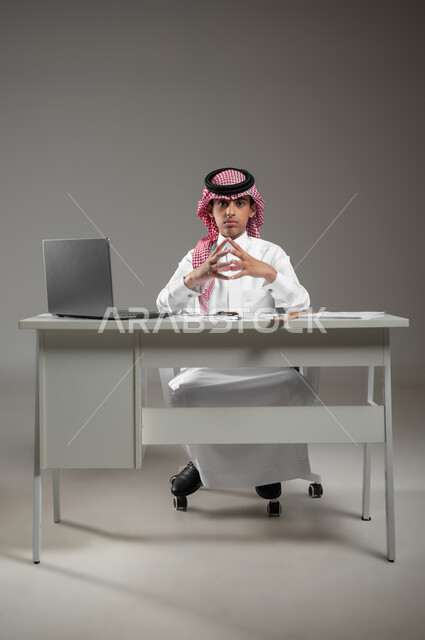 استعمال الأدوات والمعدات الهندسية، اكتساب المعرفة والخبرة بإيماءات الجدية والدقة، إستخدام الأجهزة التقنية الحديثة في المجالات العلمية، بورتريه لولد عربي خليجي سعودي يرتدي الشماغ والثوب التقليدي يجلس امام حاسوبه يقوم برسم مخطط تصميم هندسي، خلفية رمادية،محتوى حساس، استخدام حساس