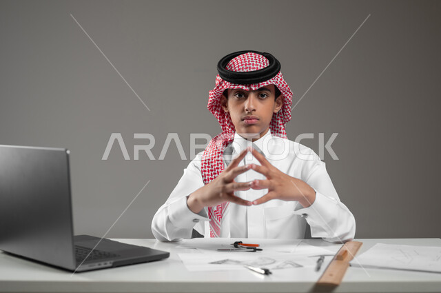 Using engineering tools and equipment, gaining knowledge and experience with gestures of seriousness and precision, using modern technical devices in scientific fields, a close-up portrait of a Saudi Gulf Arab boy wearing a shemagh and traditional dress sitting in front of his computer drawing an engineering design plan, gray background, sensitive content, sensitive use