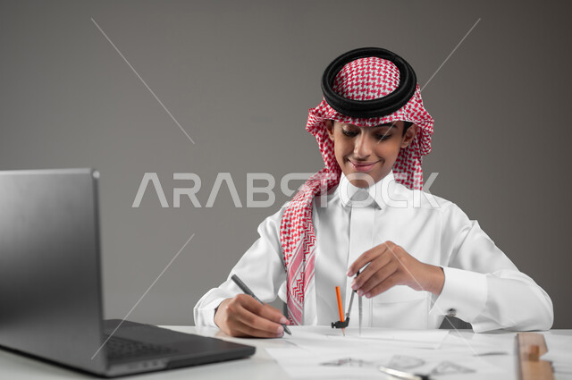إستخدام الأجهزة التقنية الحديثة في المجالات العلمية، رسم مخطط تصميم هندسي، اكتساب المعرفة والخبرة بإيماءات الدقة والتركيز، بورتريه مقرب لولد عربي خليجي سعودي يرتدي الشماغ والثوب التقليدي يجلس امام حاسوبه يقوم باستعمال الأدوات والمعدات الهندسية، خلفية رمادية، محتوى حساس، استخدام حساس