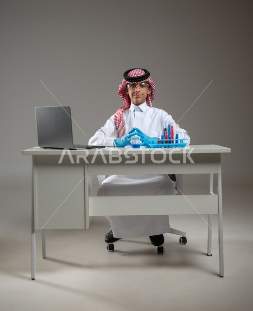 التوصل الى نتائج اثناء إجراء التفاعلات الكيميائية، التجارب والدراسات في بيئة عملية متطورة, التطوير والبحث باستخدام التكنولوجيا, بورتريه لطالب عربي خليجي سعودي يرتدي الزي التقليدي يضع نظارة الوقاية يجلس امام الحاسوب المحمول بتعابير الفخر والسرور, خلفية رمادية، محتوى حساس، استخدام حساس