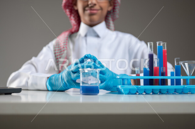 التوصل الى نتائج اثناء إجراء التفاعلات الكيميائية، التجارب والدراسات في بيئة عملية متطورة, التطوير والبحث باستخدام التكنولوجيا, بورتريه مقرب لطالب عربي خليجي سعودي يرتدي الزي التقليدي يضع نظارة الوقاية يجلس امام الحاسوب المحمول بتعابير الفخر, خلفية رمادية، استخدام حساس، محتوى حساس