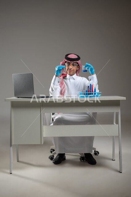 إجراء التفاعلات الكيميائية والتطوير والبحث, الدراسات والتجارب في بيئة عملية متطورة, بورتريه لطالب عربي خليجي سعودي يرتدي الزي التقليدي يضع نظارة الوقاية يجلس امام الحاسوب المحمول يقوم بتجربة علمية, اكتساب المعرفة والخبرة بإيماءات الدقة والتركيز، خلفية رمادية، محتوى حساس، استخدام حساس
