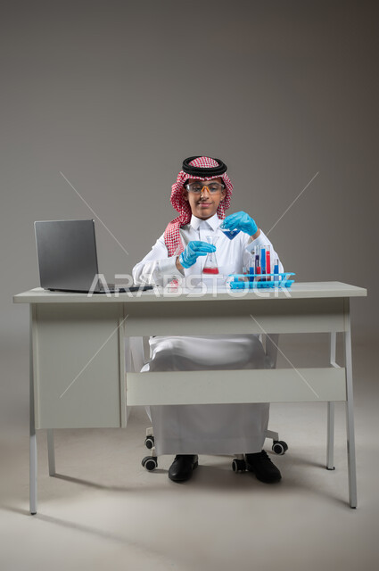 التطوير والبحث وإجراء التفاعلات الكيميائية, الدراسات والتجارب في بيئة عملية متطورة, بورتريه لطالب عربي خليجي سعودي يرتدي الزي التقليدي يضع نظارة الوقاية يجلس امام الحاسوب المحمول يقوم بتجربة علمية, اكتساب المعرفة والخبرة بإيماءات الدقة والتركيز، خلفية رمادية، محتوى حساس، استخدام حساس