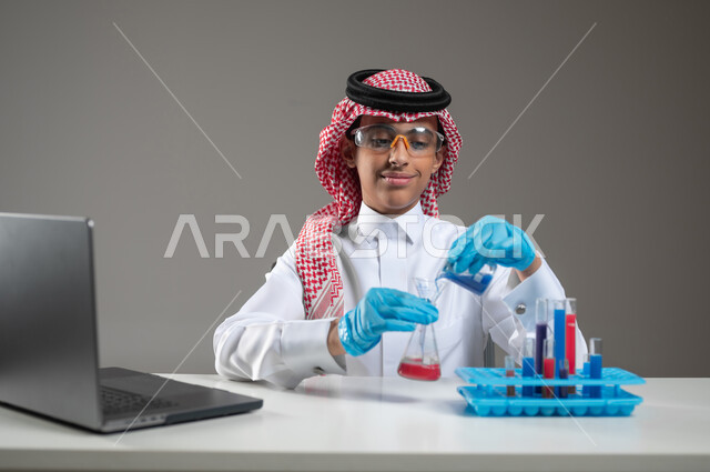 Acquiring knowledge and experience with gestures of precision and concentration, development, research, and conducting chemical reactions, studies and experiments in an advanced practical environment, close-up portrait of a Saudi Gulf Arab student wearing traditional clothing, wearing goggles, sitting in front of the laptop doing a scientific experiment, gray background, sensitive use, sensitive content