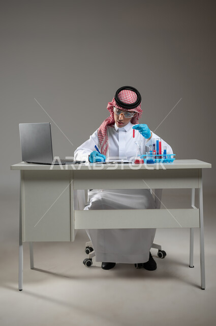 التطوير والبحث وإجراء التفاعلات الكيميائية, الدراسات والتجارب في بيئة عملية متطورة, بورتريه لطالب عربي خليجي سعودي يرتدي الزي التقليدي يضع نظارة الوقاية يجلس امام الحاسوب المحمول يقوم بتجربة علمية, اكتساب المعرفة والخبرة بإيماءات الدقة والتركيز، خلفية رمادية، محتوى حساس، استخدام حساس