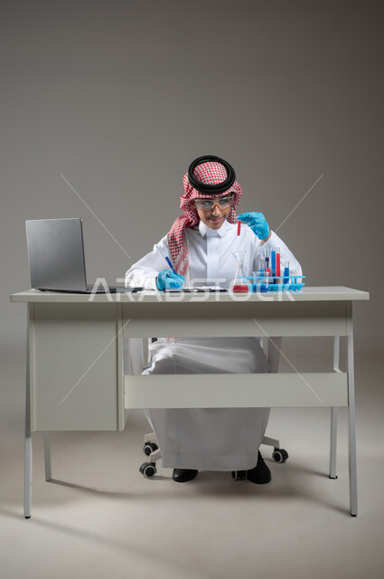 البحث والتطوير وإجراء التفاعلات الكيميائية, الدراسات والتجارب في بيئة عملية متطورة, بورتريه لطالب عربي خليجي سعودي يرتدي الزي التقليدي يضع نظارة الوقاية يجلس امام الحاسوب المحمول يقوم بتجربة علمية, اكتساب المعرفة والخبرة بإيماءات الدقة والتركيز، خلفية رمادية، محتوى حساس، استخدام حساس