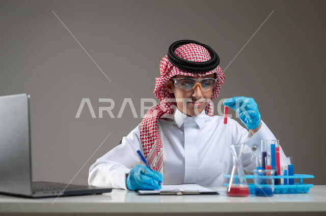 اكتساب المعرفة والخبرة بإيماءات الدقة والتركيز، التطوير والبحث وإجراء التفاعلات الكيميائية, الدراسات والتجارب في بيئة عملية متطورة, بورتريه مقرب لطالب عربي خليجي سعودي يرتدي الزي التقليدي يضع نظارة الوقاية يجلس امام الحاسوب المحمول يقوم بتجربة علمية, خلفية رمادية، محتوى حساس، استخدام حساس