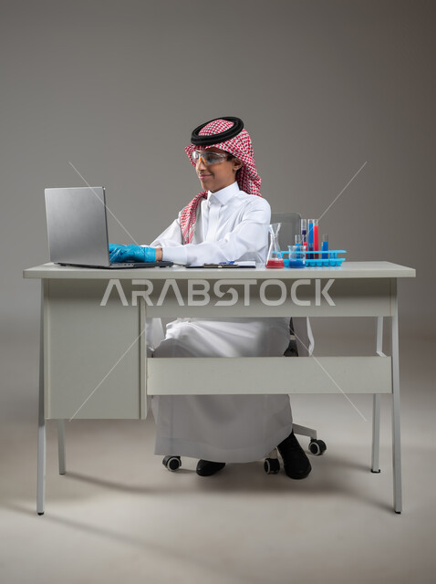 البحث والتطوير وإجراء التفاعلات الكيميائية, الدراسات والتجارب في بيئة عملية متطورة, بورتريه لطالب عربي خليجي سعودي يرتدي الزي التقليدي يضع نظارة الوقاية يجلس امام الحاسوب المحمول يقوم بتجربة علمية, اكتساب المعرفة والخبرة بإيماءات الدقة والتركيز، خلفية رمادية، محتوى حساس، استخدام حساس