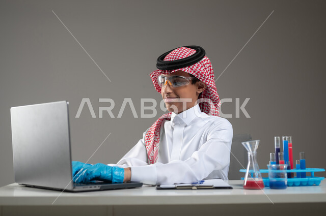 اكتساب المعرفة والخبرة بإيماءات الدقة والتركيز، التطوير والبحث وإجراء التفاعلات الكيميائية, الدراسات والتجارب في بيئة عملية متطورة, بورتريه مقرب لطالب عربي خليجي سعودي يرتدي الزي التقليدي يضع نظارة الوقاية يجلس امام الحاسوب المحمول يقوم بتجربة علمية, خلفية رمادية، محتوى حساس، استخدام حساس