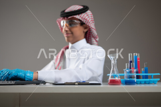 Studies and experiments in an advanced practical environment, gaining knowledge and experience with gestures of precision and concentration, development, research and conducting chemical reactions, close-up portrait of a Saudi Gulf Arab student wearing traditional clothing and wearing goggles sitting in front of the laptop doing a scientific experiment, gray background, sensitive use, sensitive content