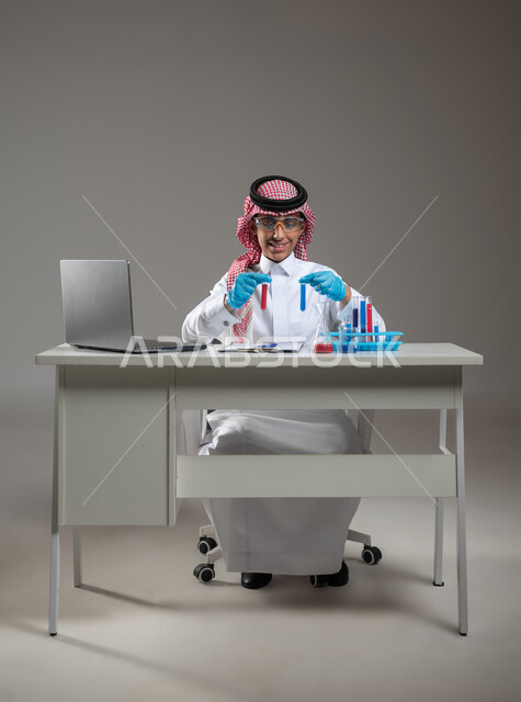 التجارب والدراسات في بيئة عملية متطورة, اكتساب المعرفة والخبرة بإيماءات الدقة والتركيز، التطوير والبحث وإجراء التفاعلات الكيميائية, بورتريه لطالب عربي خليجي سعودي يرتدي الزي التقليدي يضع نظارة الوقاية يجلس امام الحاسوب المحمول يقوم بتجربة علمية, خلفية رمادية، استخدام حساس، محتوى حساس