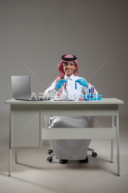 Experiments and studies in an advanced practical environment, gaining knowledge and experience with gestures of precision and concentration, development, research, and conducting chemical reactions, portrait of a Saudi Arabian Gulf student wearing traditional clothing, wearing goggles, sitting in front of the laptop doing a scientific experiment, gray background, sensitive use, sensitive content