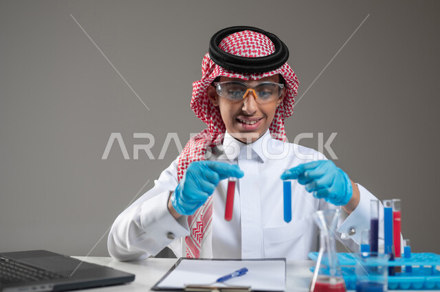 الدراسات والتجارب في بيئة عملية متطورة, اكتساب المعرفة والخبرة بإيماءات الدقة والتركيز، التطوير والبحث وإجراء التفاعلات الكيميائية, بورتريه مقرب لطالب عربي خليجي سعودي يرتدي الزي التقليدي يضع نظارة الوقاية يجلس امام الحاسوب المحمول يقوم بتجربة علمية, خلفية رمادية، استخدام حساس، محتوى حساس
