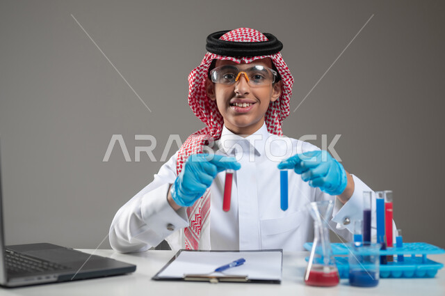 الدراسات والتجارب في بيئة عملية متطورة, اكتساب المعرفة والخبرة بإيماءات الدقة والتركيز، التطوير والبحث وإجراء التفاعلات الكيميائية, بورتريه مقرب لطالب عربي خليجي سعودي يرتدي الزي التقليدي يضع نظارة الوقاية يجلس امام الحاسوب المحمول يقوم بتجربة علمية, خلفية رمادية، استخدام حساس، محتوى حساس