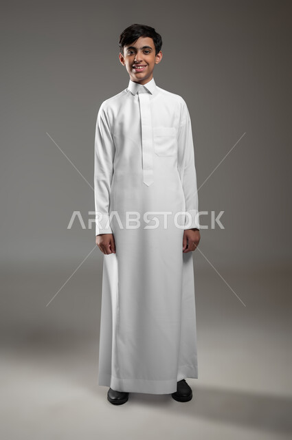 مفهوم والأناقه والاهتمام بالمظهر الخارجي، بورتريه لولد عربي خليجي سعودي مبتسم يرتدي الثوب التقليدي يقف باستقامة وينظر إلى الكاميرا يإيماءات السرور، التقاط صور شخصية للوثائق الرسمية، صورة الجسم بالطول الكامل، خلفية رمادية، استخدام حساس، محتوى حساس