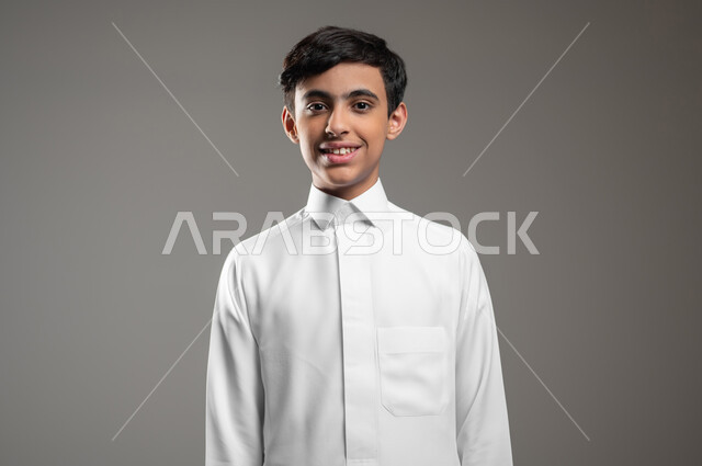 Concept, elegance and attention to external appearance, close-up portrait of a smiling Saudi Arabian Gulf boy wearing a traditional dress, standing straight and looking at the camera with happy gestures, taking personal photos for official documents, gray background, sensitive use, sensitive content