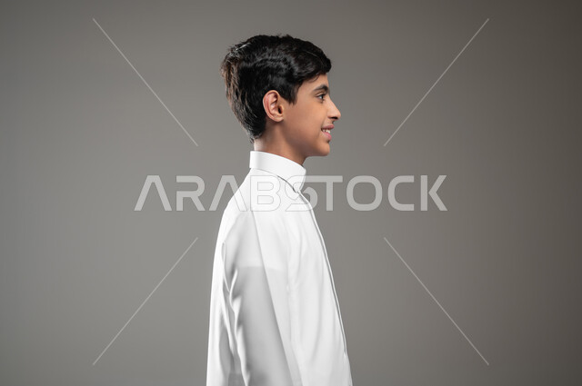 الأناقه والاهتمام بالمظهر الخارجي، بورتريه من الجانب لولد عربي خليجي سعودي مبتسم يرتدي الثوب التقليدي يقف باستقامة وينظر إلى الكاميرا يإيماءات السرور، التقاط صور شخصية للوثائق الرسمية، صورة الجسم بالطول الكامل، خلفية رمادية، محتوى حساس، استخدام حساس