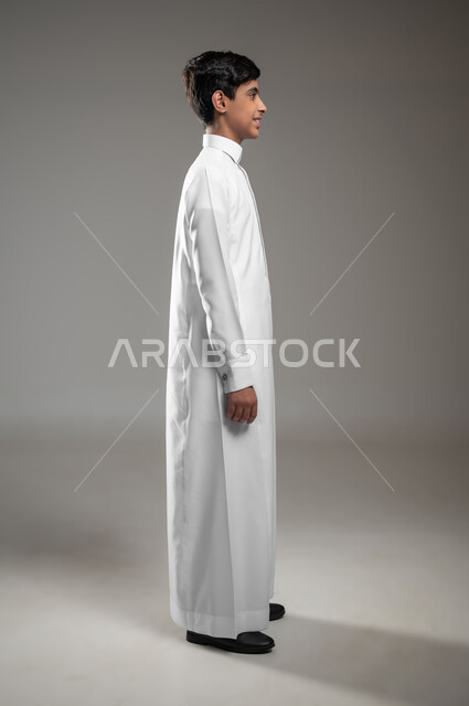 الأناقه والاهتمام بالمظهر الخارجي، بورتريه مقرب من الجانب لولد عربي خليجي سعودي مبتسم يرتدي الثوب التقليدي يقف باستقامة وينظر إلى الكاميرا يإيماءات السرور، التقاط صور شخصية للوثائق الرسمية، خلفية رمادية، محتوى حساس، استخدام حساس