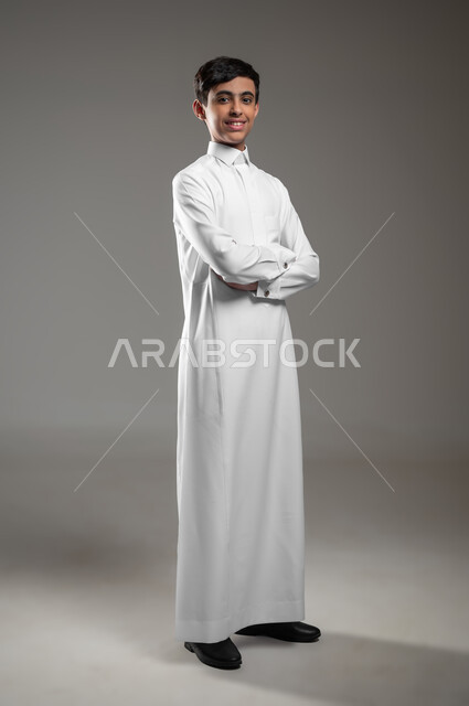 Standing with crossed hands with an expression of self-confidence, portrait of a Saudi Arabian Gulf boy wearing a traditional dress looking at the camera with happy gestures, taking personal photos for official documents, elegance and attention to external appearance, full-length body photo, gray background, sensitive use, sensitive content