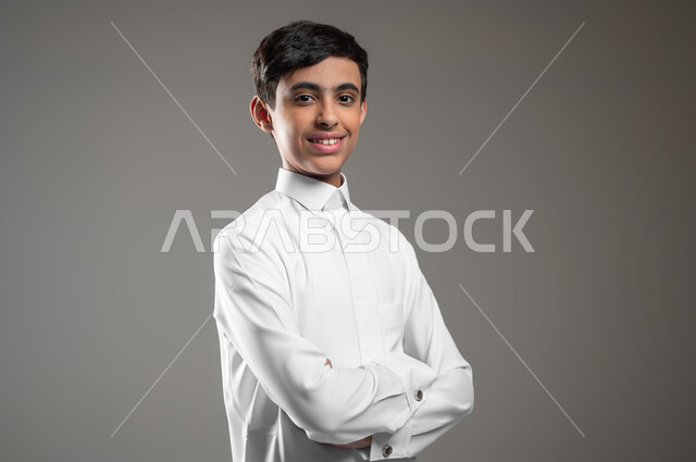 الوقوف بأيدي متقاطعة بتعابير الثقة بالنفس، بورتريه مقرب لولد عربي خليجي سعودي يرتدي الثوب التقليدي ينظر إلى الكاميرا يإيماءات السرور، التقاط صور شخصية للوثائق الرسمية، الأناقه والاهتمام بالمظهر الخارجي، خلفية رمادية، محتوى حساس، استخدام حساس